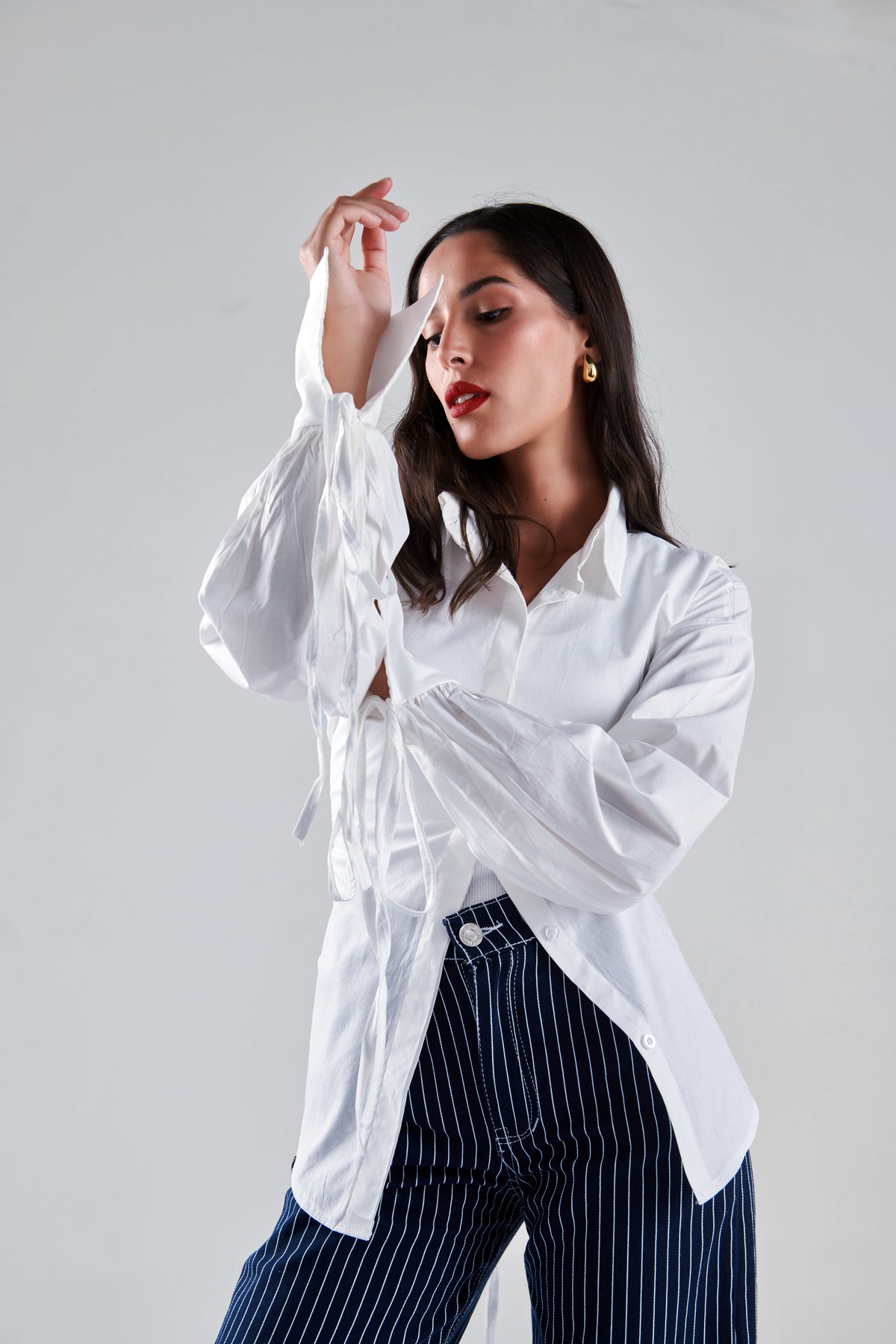 A woman posing in a white long-sleeve button-up shirt with sheer tie details at the cuffs and navy blue pinstriped trousers, set against a plain light gray studio background.