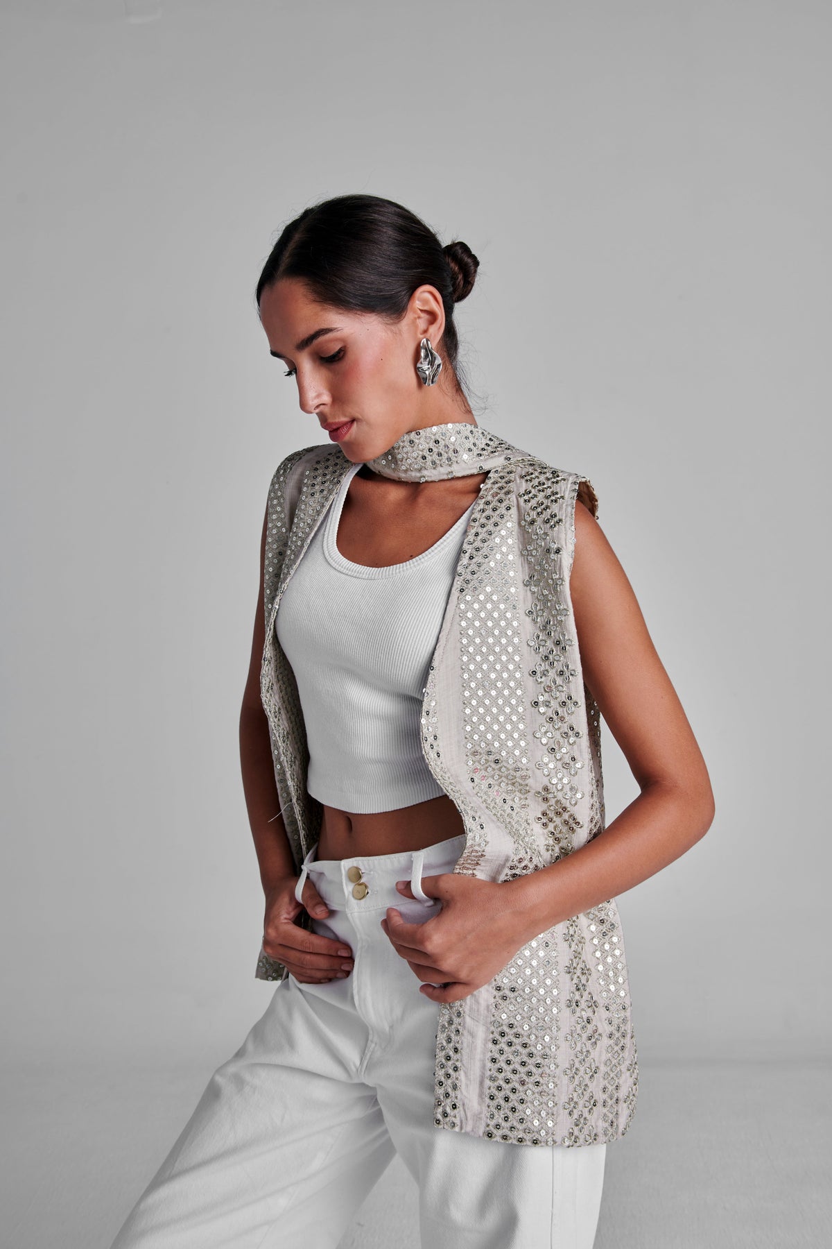 A woman models a sleeveless, open-front vest covered in small silver studs over a white crop top and white pants, photographed against a plain light gray studio background. #Beige