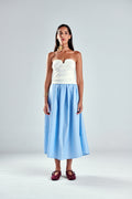 A model stands against a light gray studio background wearing a strapless white top and a light blue and white vertically striped midi skirt, paired with dark red strappy sandals.