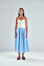 A model stands against a light gray studio background wearing a strapless white top and a light blue and white vertically striped midi skirt, paired with dark red strappy sandals.