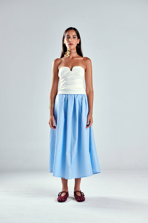 A model stands against a light gray studio background wearing a strapless white top and a light blue and white vertically striped midi skirt, paired with dark red strappy sandals.