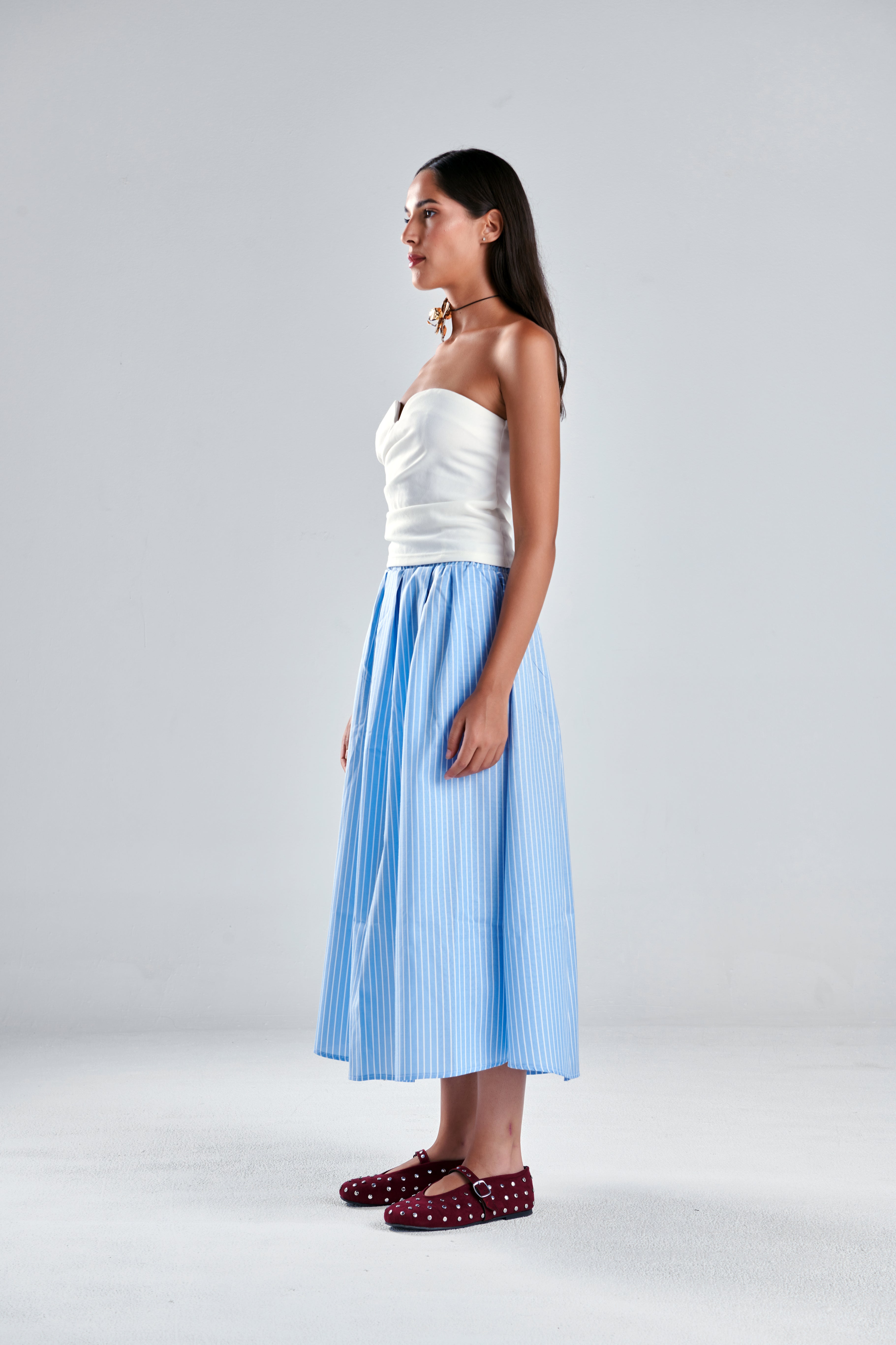 A woman stands in profile wearing a white strapless top and a light blue pleated midi skirt, paired with burgundy polka-dot flats, against a plain light gray studio background.