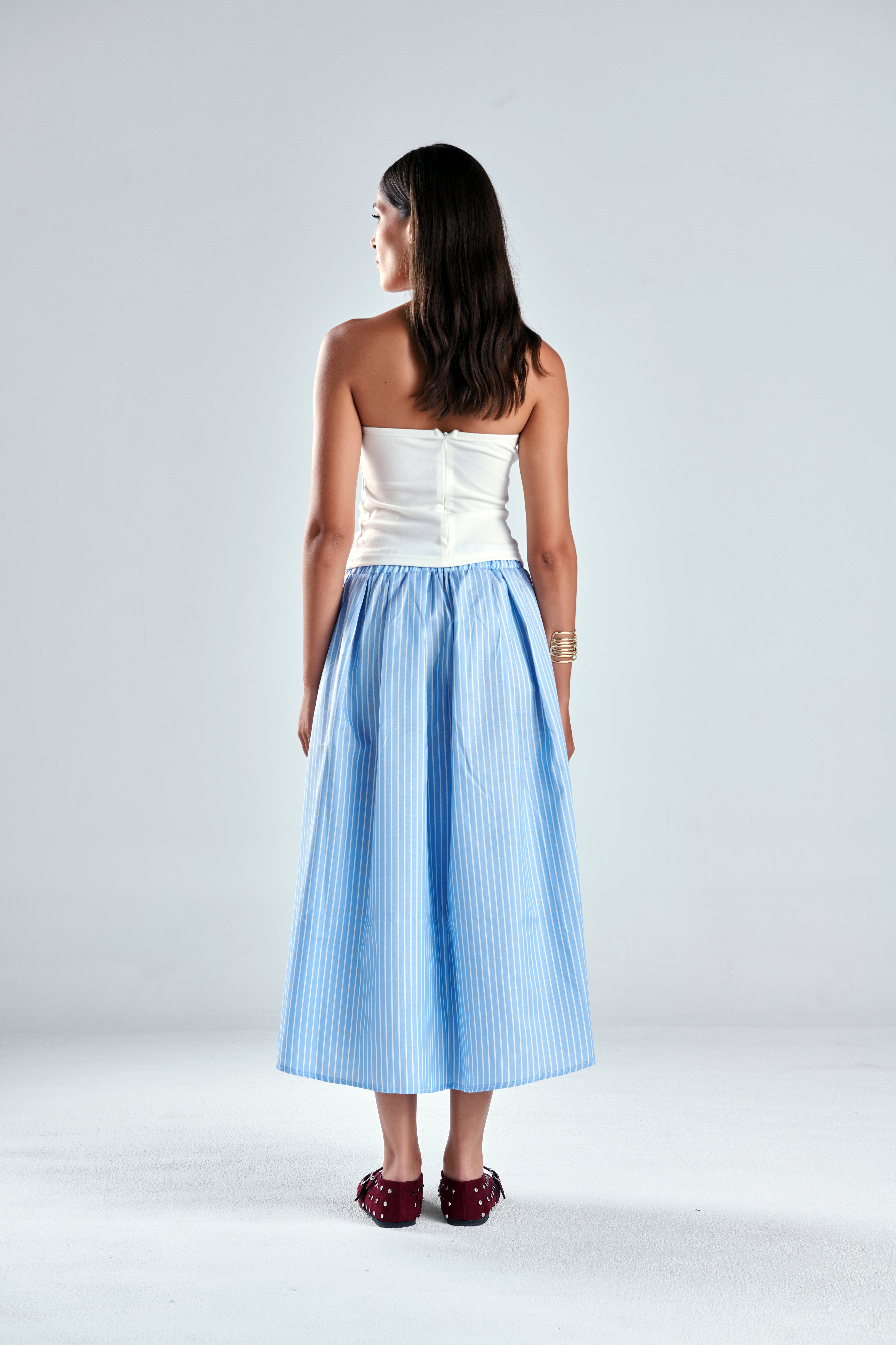 A woman stands with her back to the camera wearing a strapless white top and a light blue and white vertically striped midi skirt, paired with dark red shoes, against a light gray studio background.