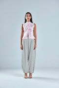 A woman stands against a light gray studio background wearing a sleeveless light pink top with a large bow at the neckline and loose white pants with black polka dots.