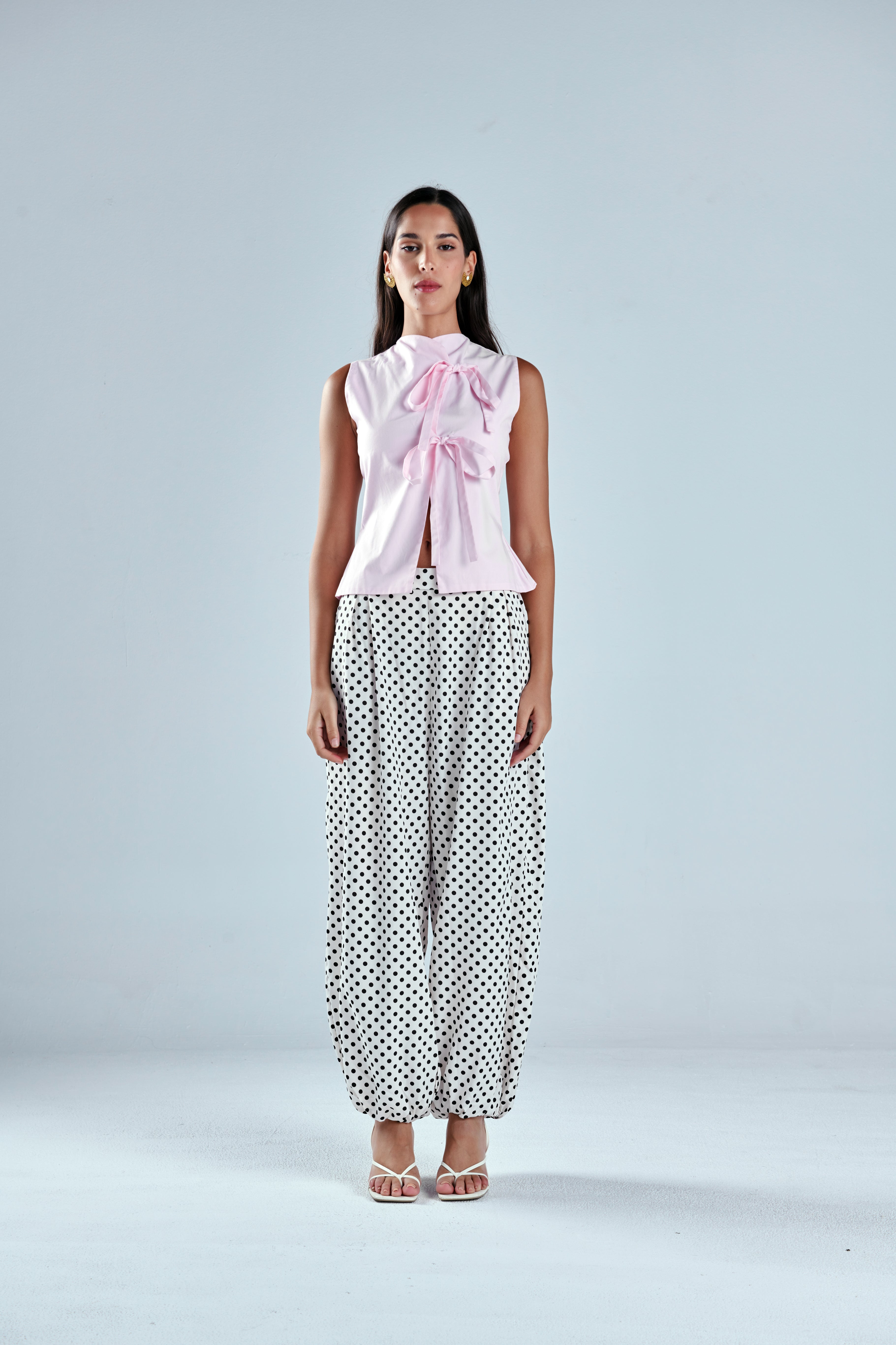 A woman stands against a light gray studio background wearing a sleeveless light pink top with a large bow at the neckline and loose white pants with black polka dots.