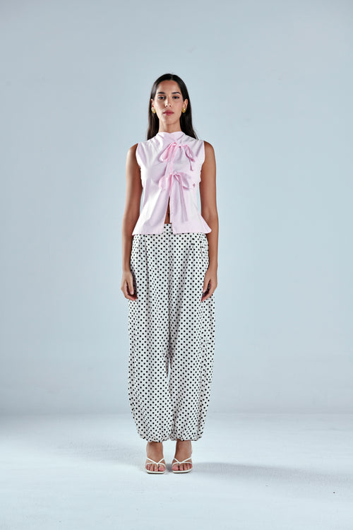 A woman stands against a light gray studio background wearing a sleeveless light pink top with a large bow at the neckline and loose white pants with black polka dots.