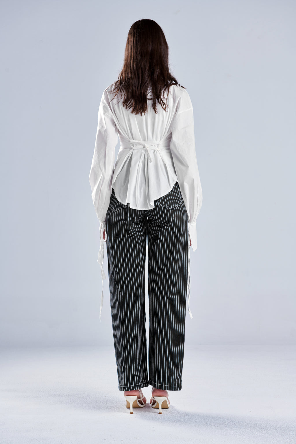 A woman shown from the back wearing a white long-sleeve blouse with a tie waist and dark gray pinstriped wide-leg trousers, standing against a light gray studio background.