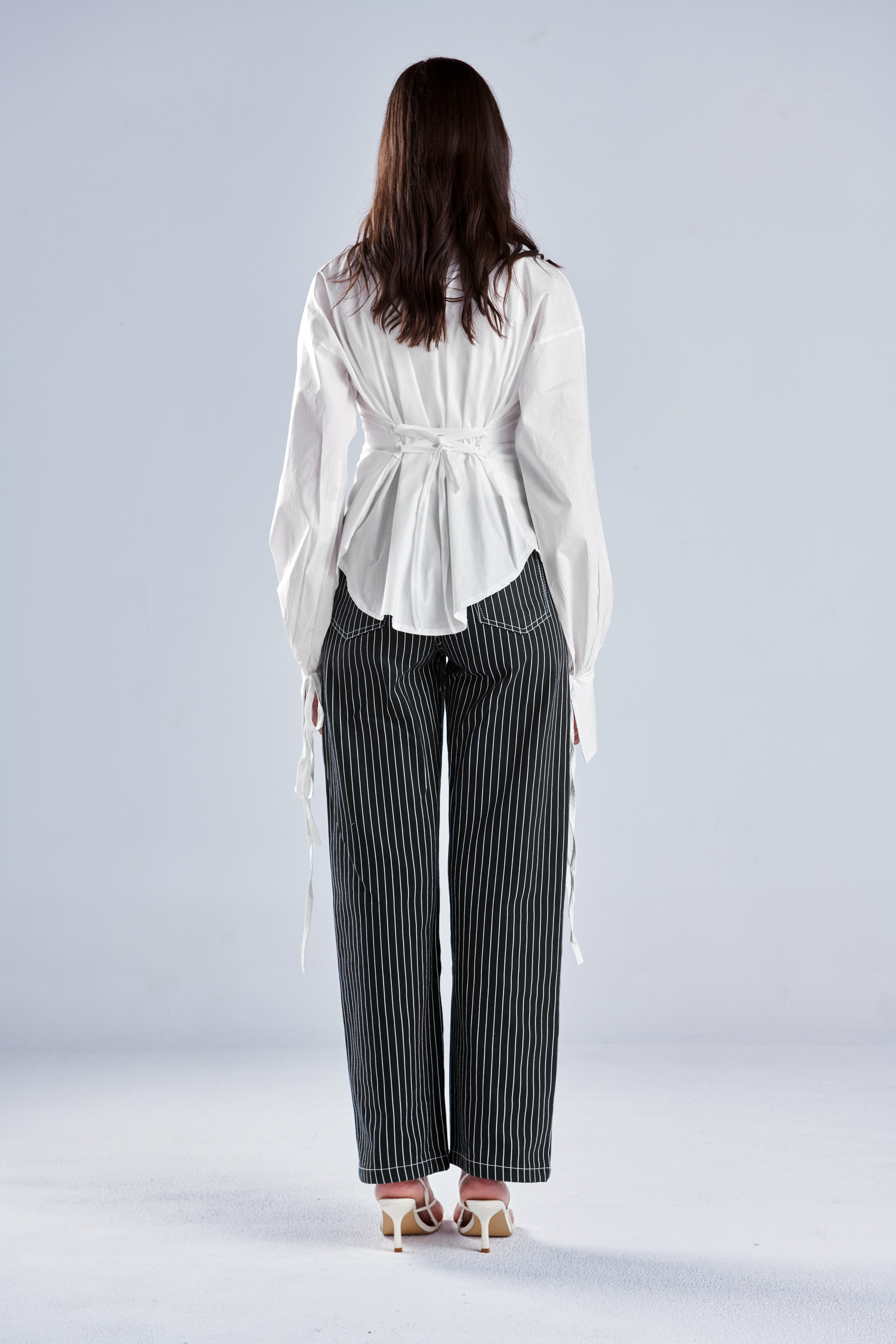 A woman shown from the back wearing a white long-sleeve blouse with a tie waist and dark gray pinstriped wide-leg trousers, standing against a light gray studio background.