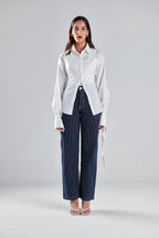 A woman stands against a light gray studio background wearing a white long-sleeve button-up shirt with gathered shoulders and side ties, paired with navy pinstriped wide-leg trousers and clear high-heel sandals.