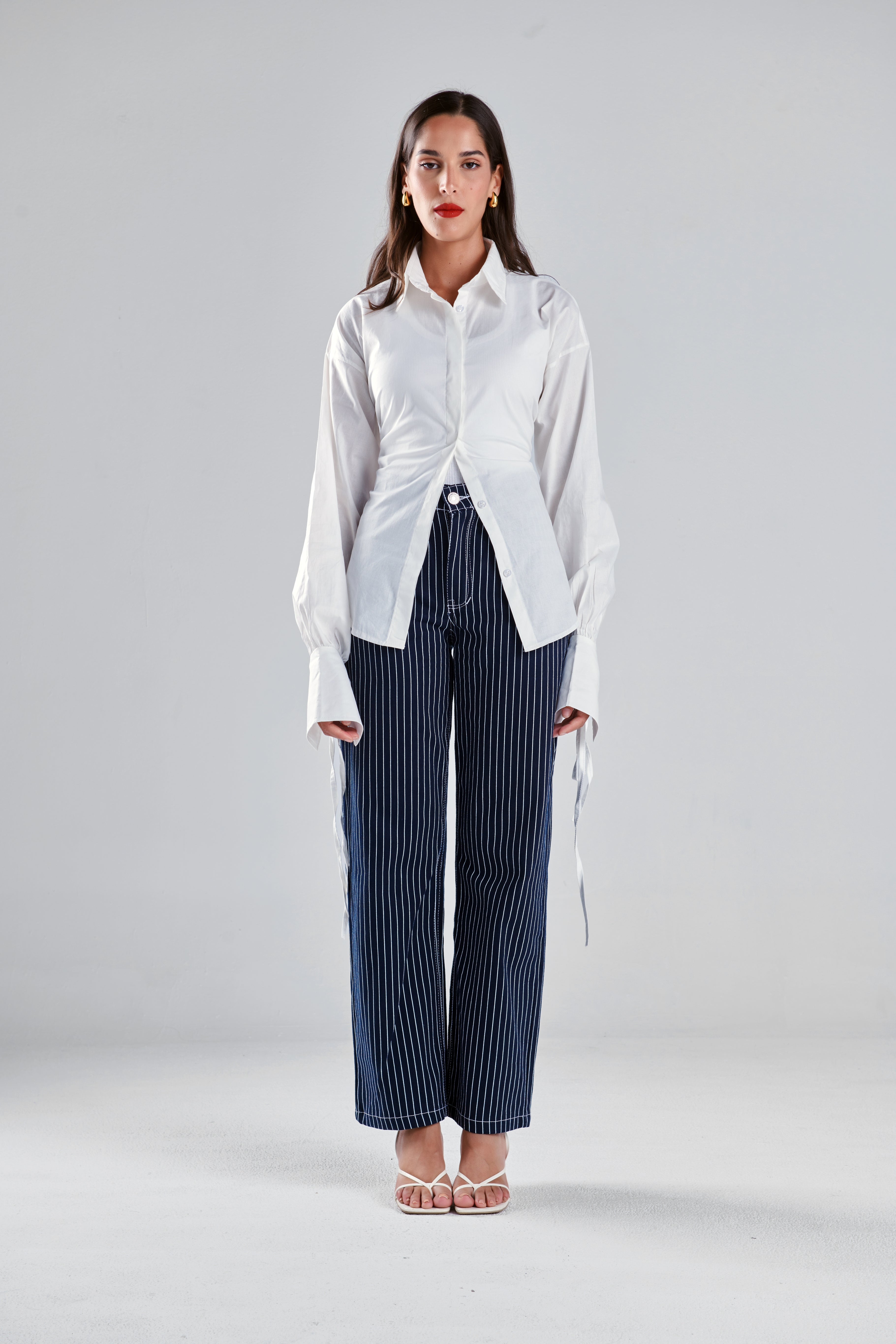 A woman stands against a light gray studio background wearing a white long-sleeve button-up shirt with gathered shoulders and side ties, paired with navy pinstriped wide-leg trousers and clear high-heel sandals.