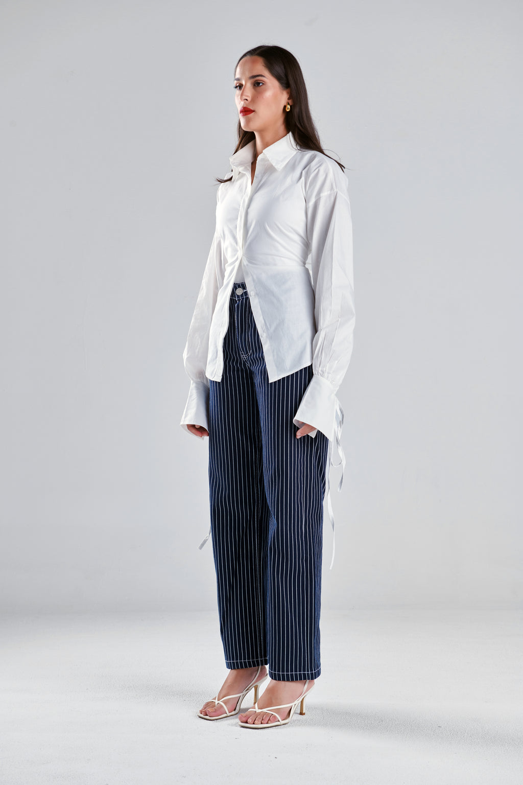 A woman modeling a white long-sleeve button-up shirt with side ties and dark blue pinstriped wide-leg trousers, standing against a light gray studio background.