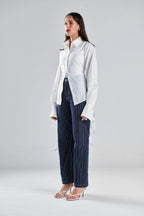 A woman modeling a white long-sleeve button-up shirt with side ties and dark blue pinstriped wide-leg trousers, standing against a light gray studio background.