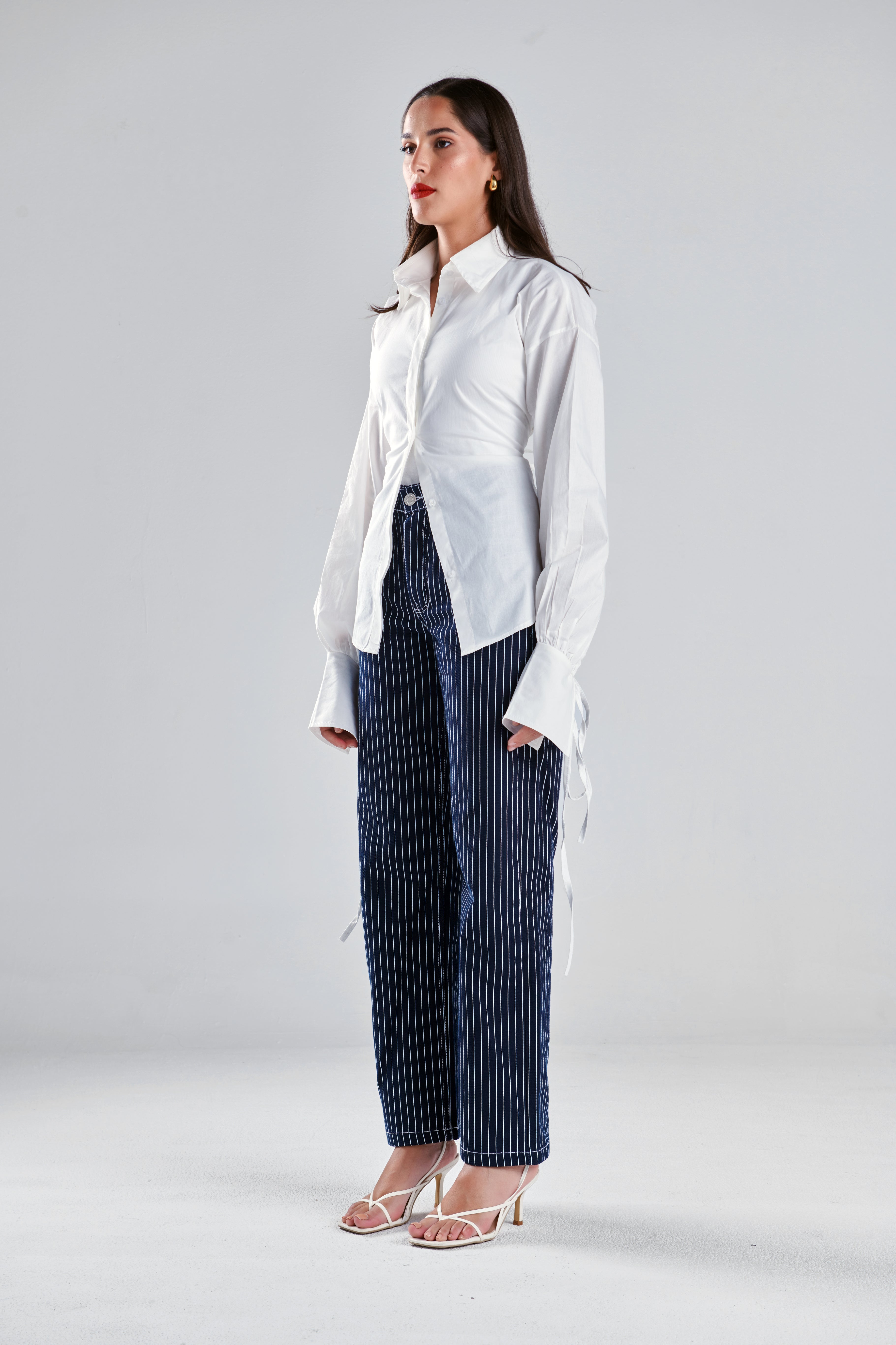 A woman modeling a white long-sleeve button-up shirt with side ties and dark blue pinstriped wide-leg trousers, standing against a light gray studio background.