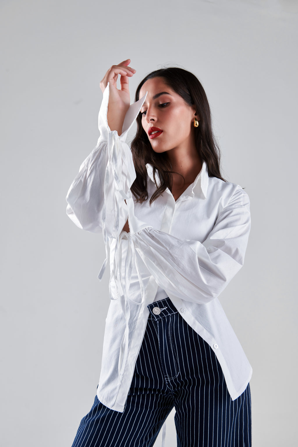 A woman posing in a white long-sleeve button-up shirt with sheer tie details at the cuffs and navy blue pinstriped trousers, set against a plain light gray studio background.