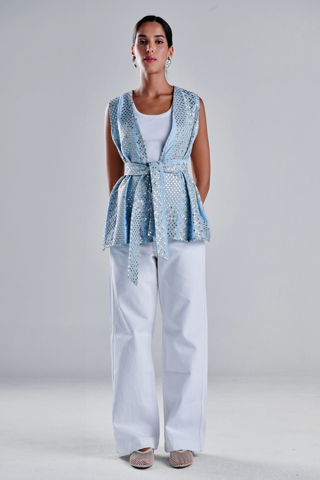 A woman stands against a light gray studio background wearing a sleeveless light blue patterned wrap-style vest over a white top and white wide-leg pants, with beige pointed flats. #Baby Blue