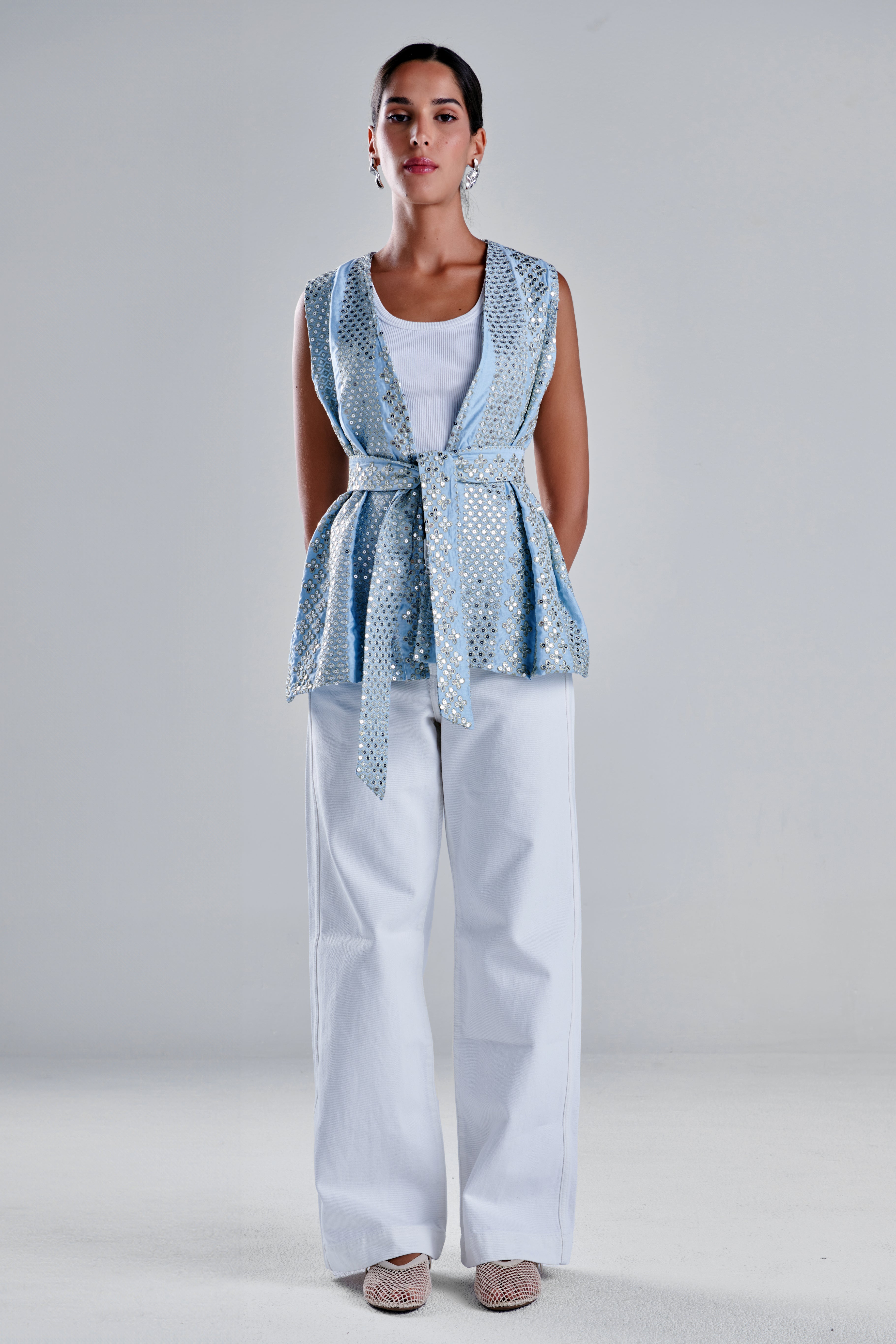 A woman stands against a light gray studio background wearing a sleeveless light blue patterned wrap-style vest over a white top and white wide-leg pants, with beige pointed flats. #Baby Blue