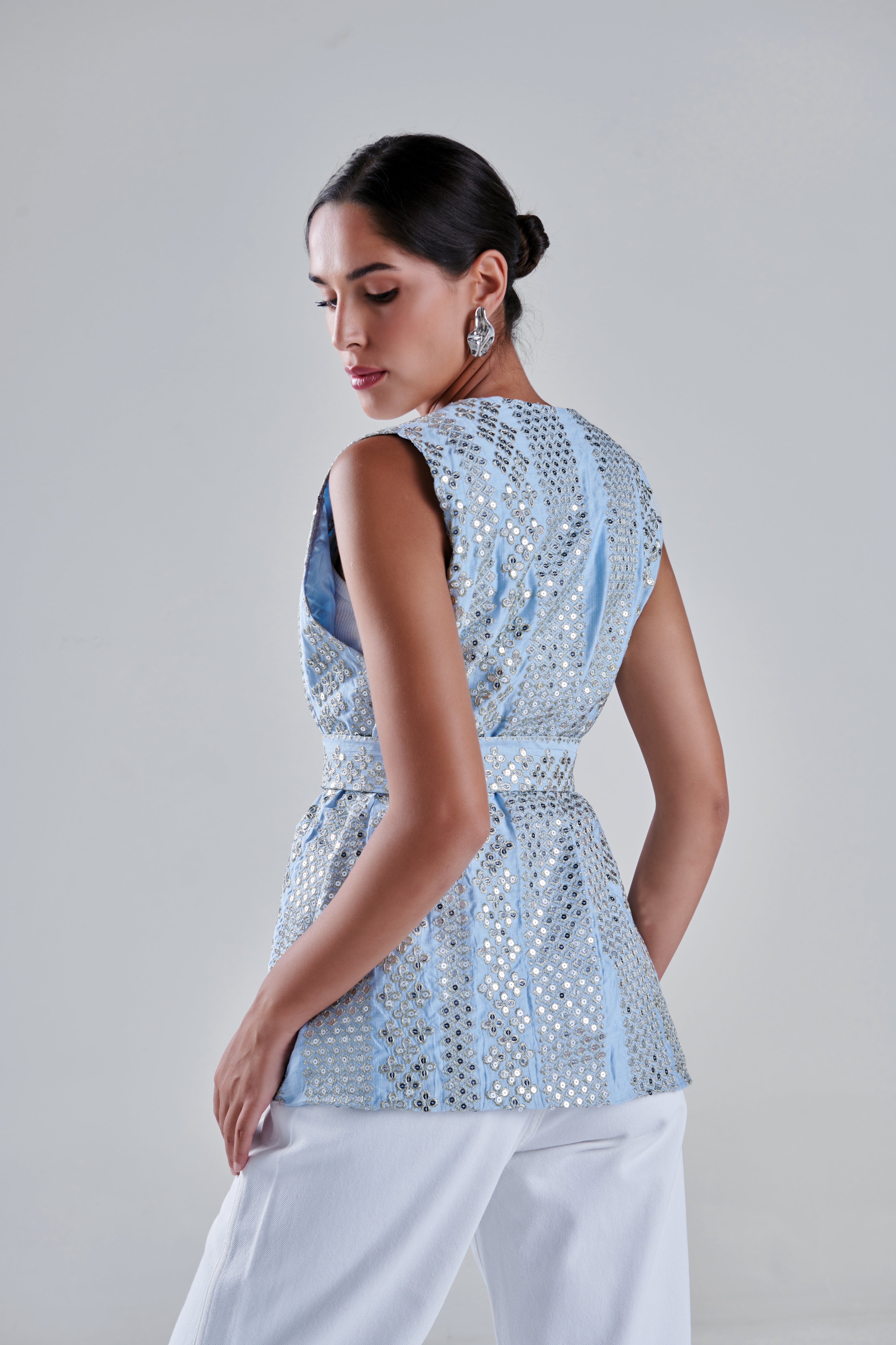 A woman shown from the back wearing a light blue sleeveless top with silver mirror work and white pants, photographed against a plain light gray studio background. #Baby Blue