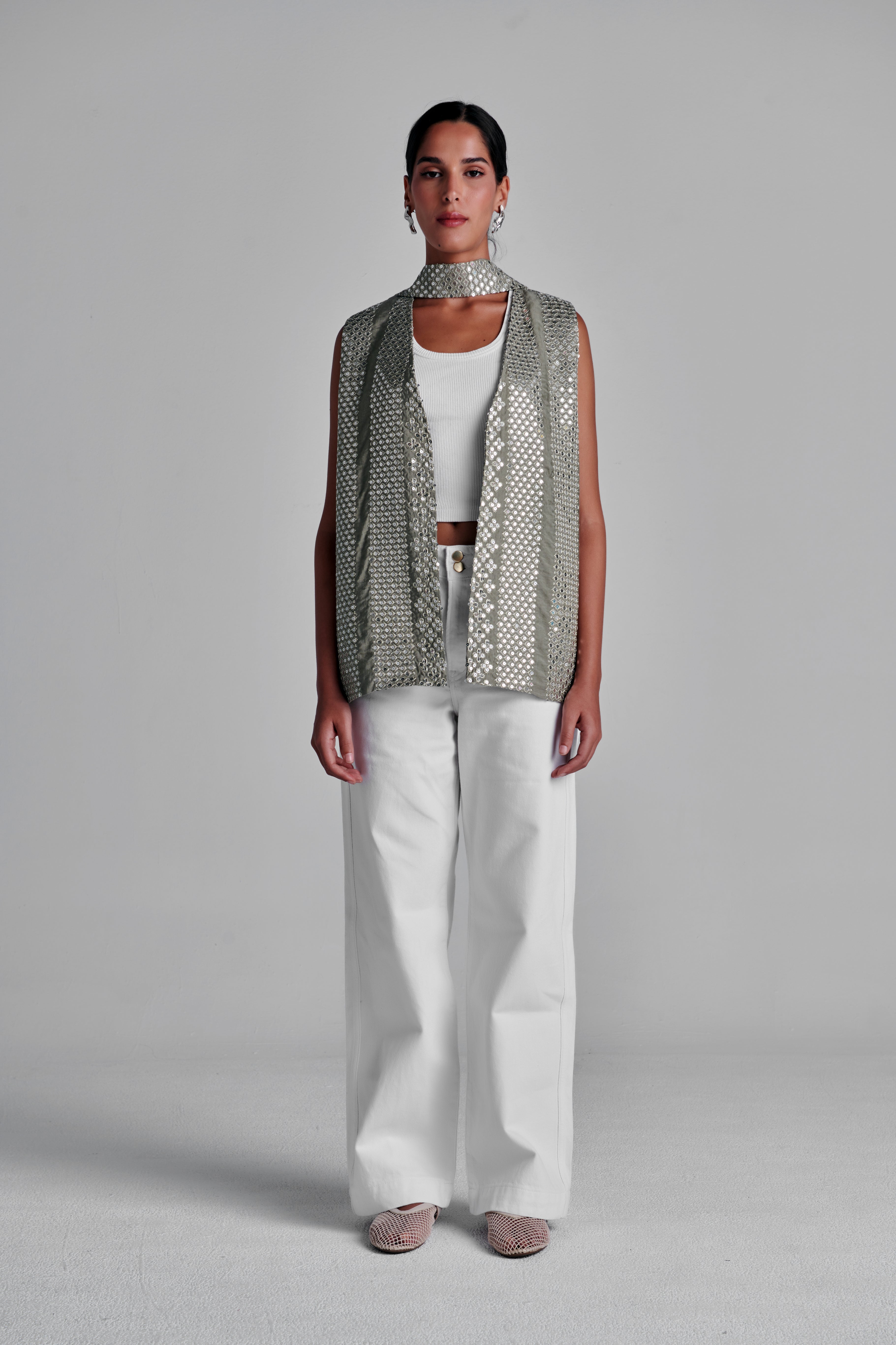 A model stands against a light gray studio background wearing a sleeveless silver sequined vest over a white top and white wide-leg pants. #Olive