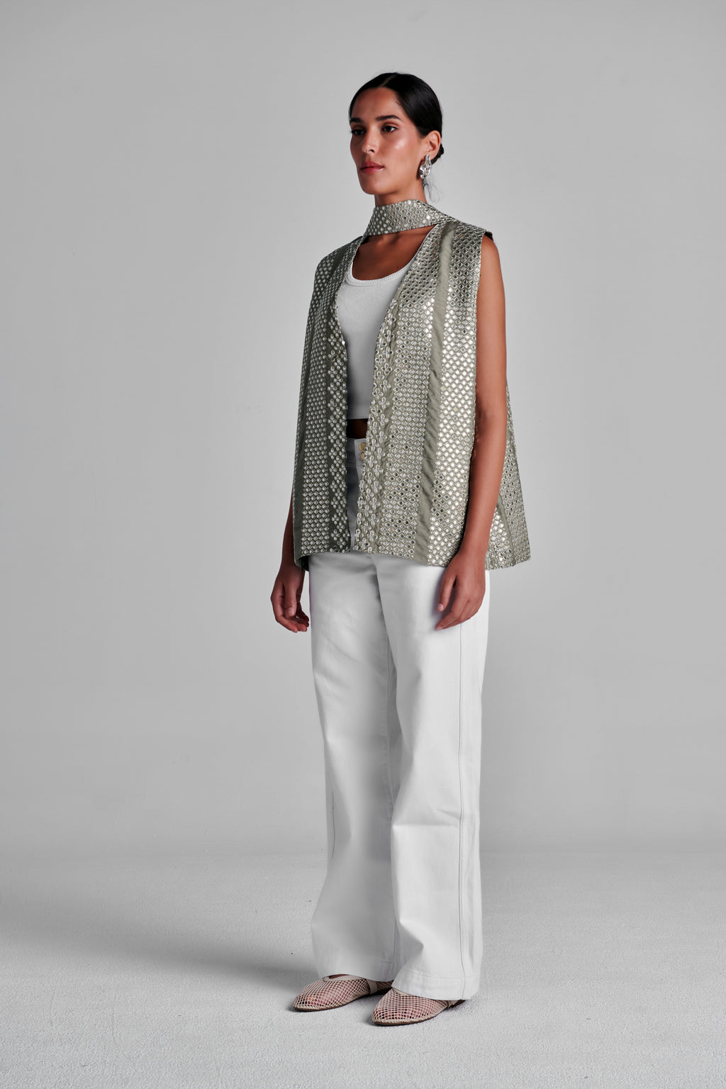 A model stands against a light gray studio background wearing a sleeveless metallic silver mesh vest with a high mock neck, white wide-leg trousers, and beige pointed flats. #Olive