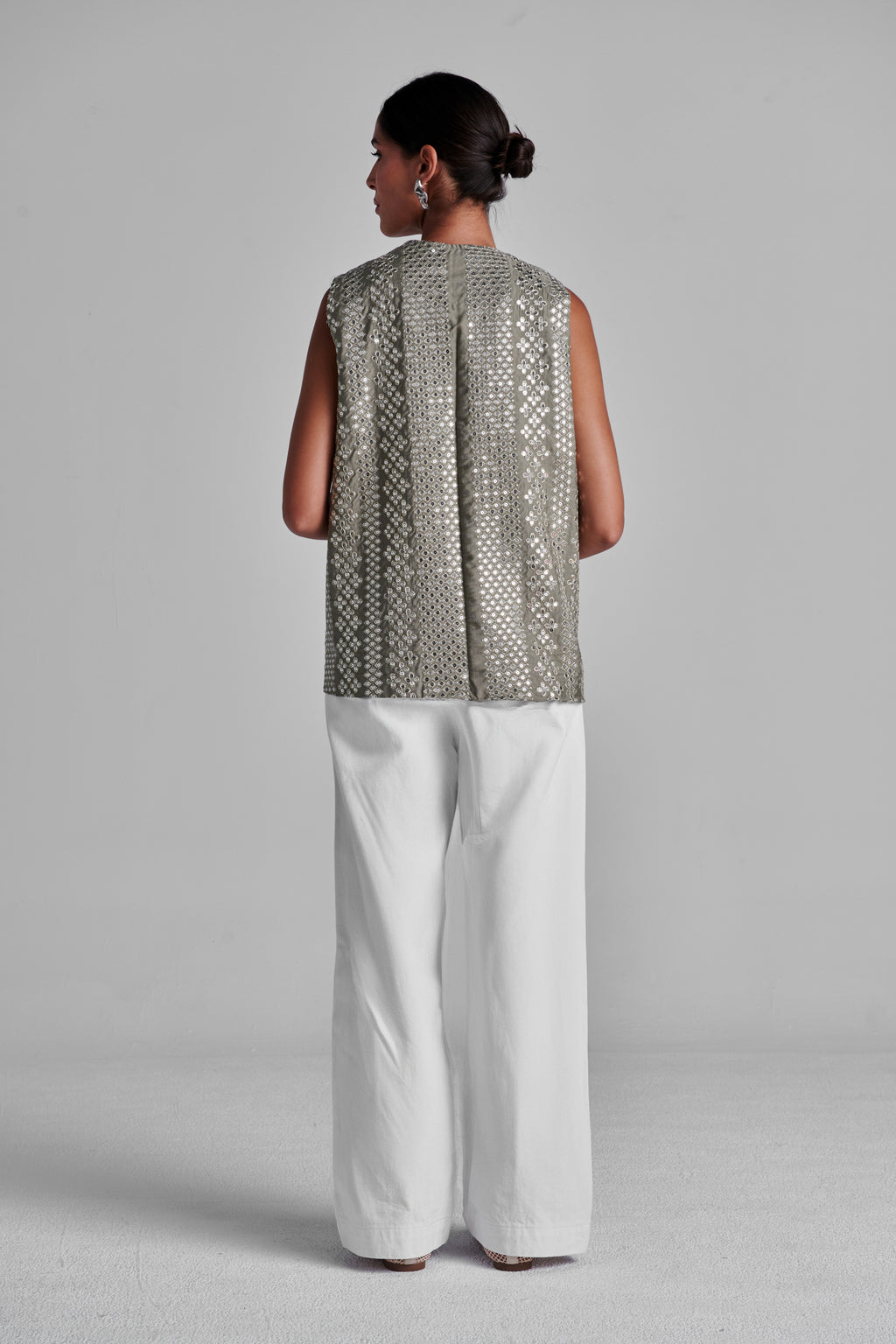 A woman is shown from the back wearing a sleeveless silver patterned top and white wide-leg trousers against a light gray studio background. #Olive