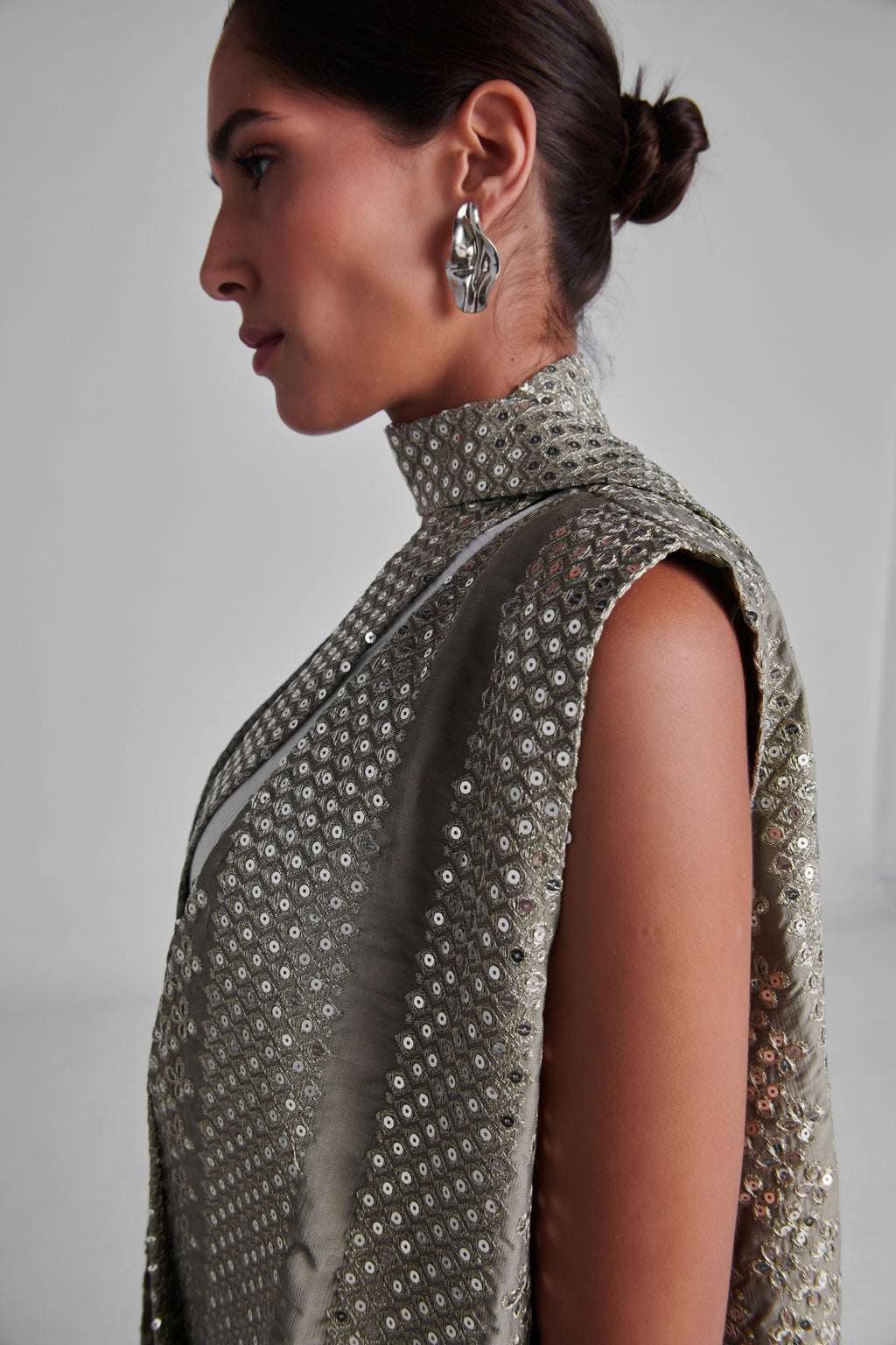 A side-profile view of a model wearing a sleeveless high-neck turtleneck dress covered in shimmering silver sequins, paired with a matching draped scarf and silver statement earrings. #Olive