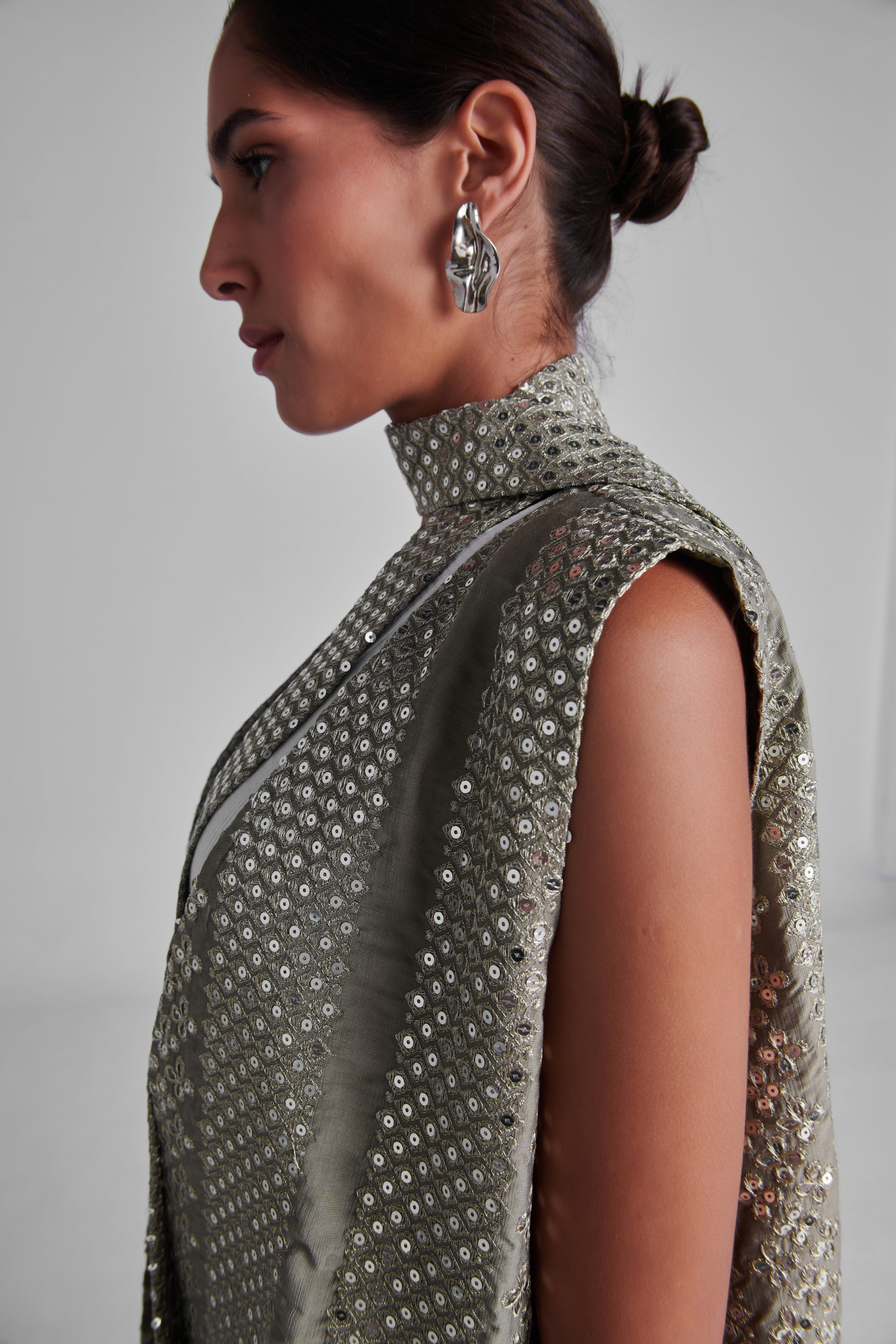 A side-profile view of a model wearing a sleeveless high-neck turtleneck dress covered in shimmering silver sequins, paired with a matching draped scarf and silver statement earrings. #Olive