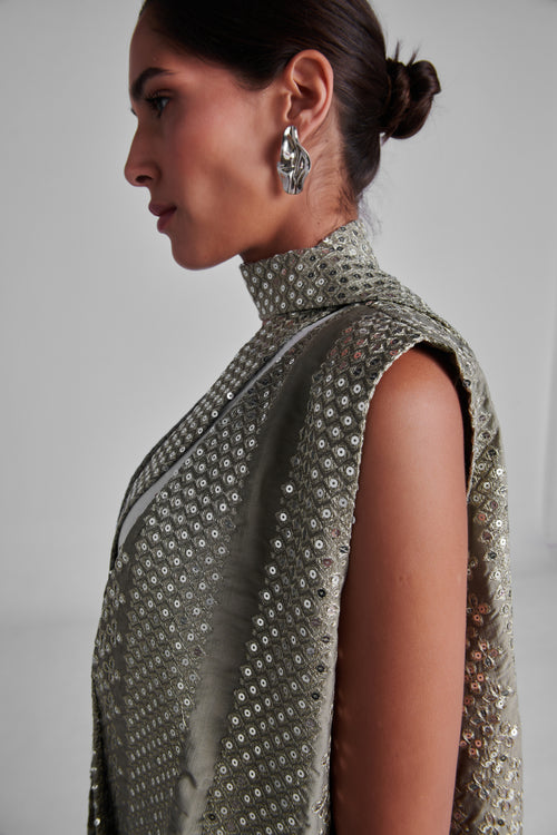 A side-profile view of a model wearing a sleeveless high-neck turtleneck dress covered in shimmering silver sequins, paired with a matching draped scarf and silver statement earrings. #Olive