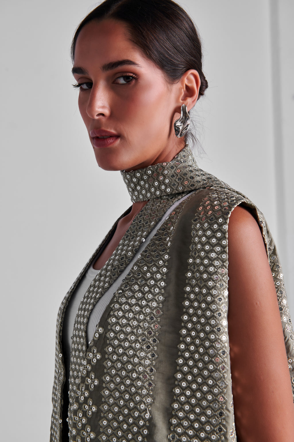A close-up of a model wearing a sleeveless high-neck top covered in shimmering silver sequins over a light gray underlayer, set against a plain light gray background. #Olive