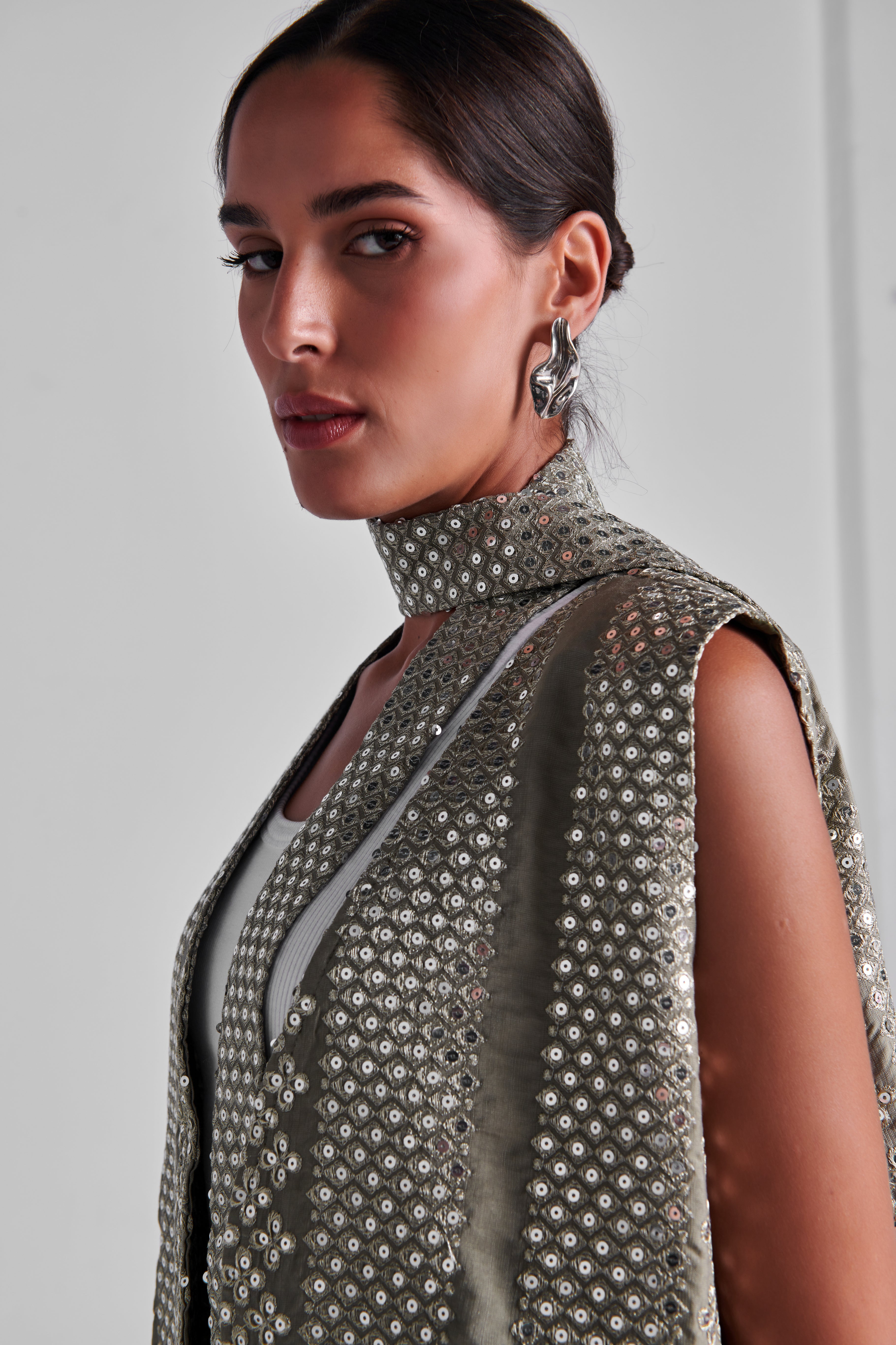 A close-up of a model wearing a sleeveless high-neck top covered in shimmering silver sequins over a light gray underlayer, set against a plain light gray background. #Olive