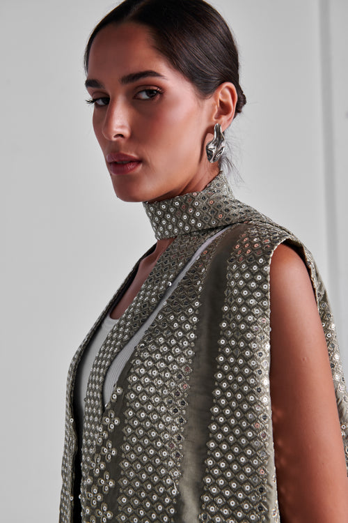 A close-up of a model wearing a sleeveless high-neck top covered in shimmering silver sequins over a light gray underlayer, set against a plain light gray background. #Olive