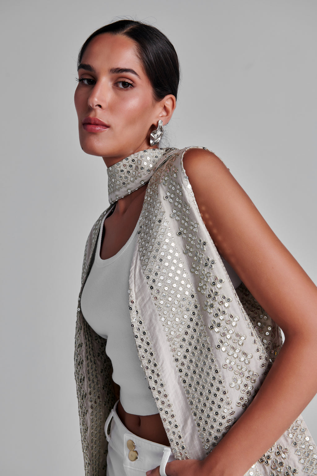 A woman wearing a sleeveless, high-neck silver embellished vest over a white crop top and white pants, photographed against a plain light gray studio background. #Beige