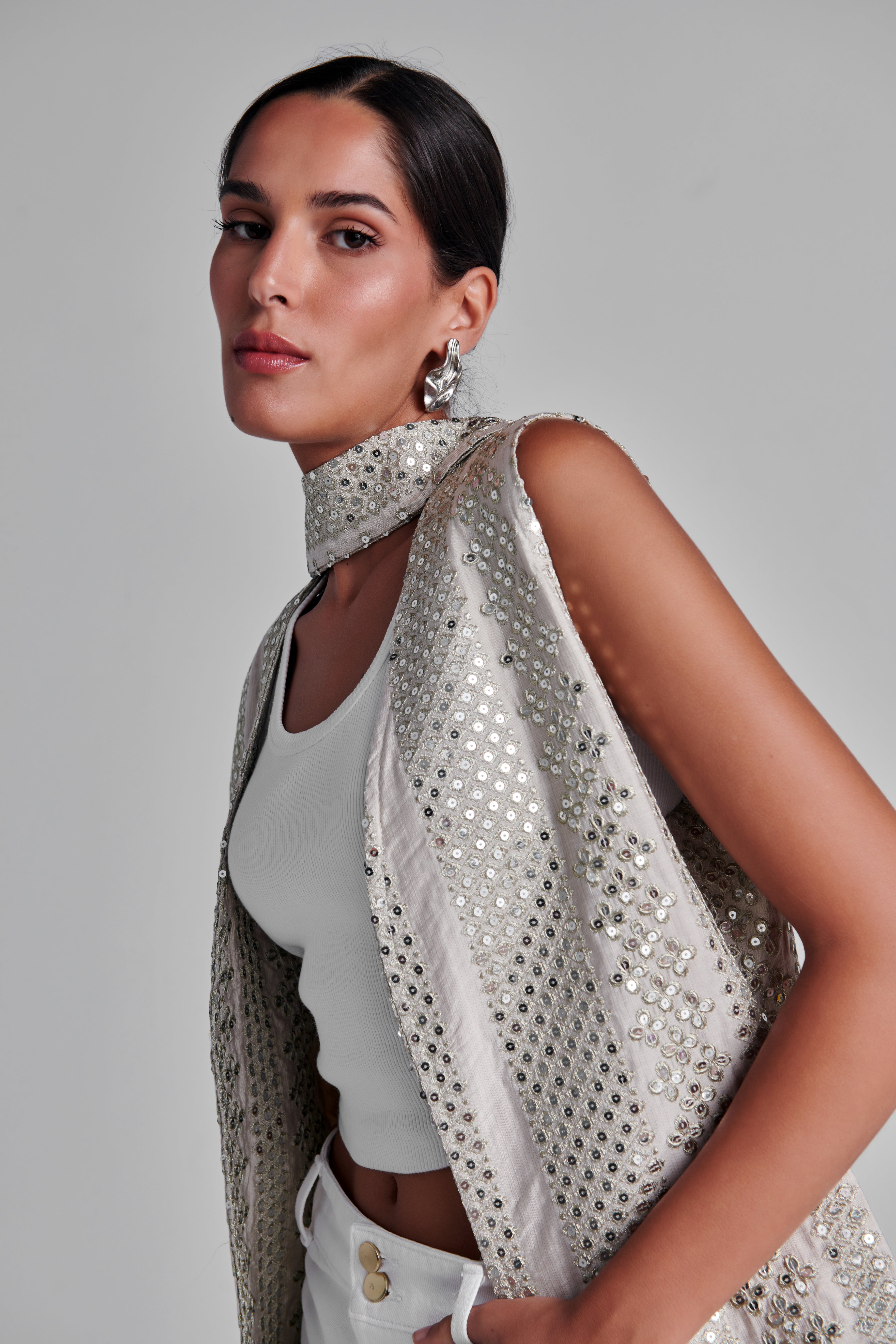 A woman wearing a sleeveless, high-neck silver embellished vest over a white crop top and white pants, photographed against a plain light gray studio background. #Beige
