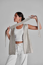 A model wearing a white crop top and white trousers, holding up a long, sleeveless, open-front vest covered in silver rhinestones over a white background. #Beige